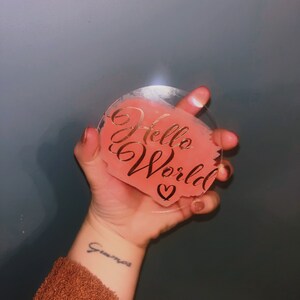May include: A clear, heart-shaped acrylic coaster with a coral-coloured centre. The words "Hello World" are written in gold script, with a small heart below. The coaster is held in a hand.