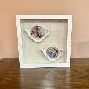 Peut inclure: Une œuvre d'art encadrée en blanc avec deux pièces en céramique en forme de théière, chacune remplie de fleurs séchées dans des tons de violet, de jaune et de rouge. Les théières sont posées sur un fond en tissu neutre.