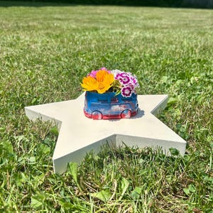 May include: A small, blue and red resin planter shaped like a vintage van, filled with a bouquet of bright orange, pink, and white flowers, resting on a flat, off-white, five-pointed star-shaped wooden base.