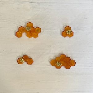 May include: Four honeycomb-shaped resin ornaments with a gold bee accent. The ornaments are different sizes and are arranged on a white wooden surface.
