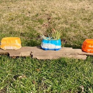 May include: Three colorful ceramic planters shaped like vintage Volkswagen vans. The planters are sitting on a wooden surface. One planter is blue and white, one is orange, and one is yellow.