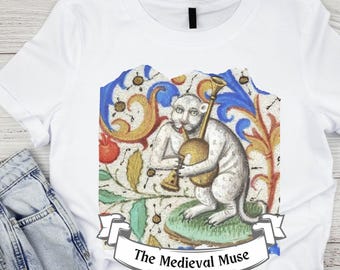 Op de afbeelding: Wit T-shirt met een middeleeuwse illustratie van een witte kat die een muziekinstrument bespeelt. De illustratie is omgeven door een kleurrijk bloemmotief. De tekst "The Medieval Muse" is onder de afbeelding gedrukt.