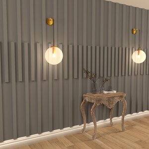 May include: A wooden console table with a carved design and a distressed white finish. Two gold wall sconces with clear glass globes illuminate the wall behind the table. The wall is painted a dark gray and features a vertical wood panel design.