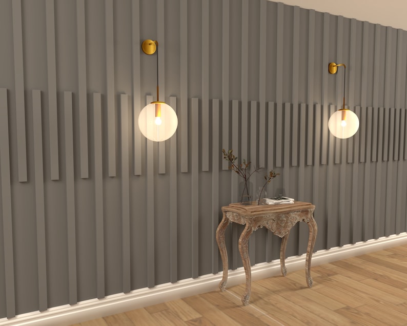 May include: A wooden console table with a carved design and a distressed white finish. Two gold wall sconces with clear glass globes illuminate the wall behind the table. The wall is painted a dark gray and features a vertical wood panel design.