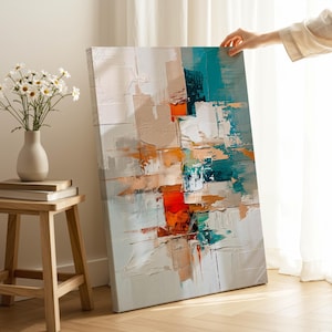 Könnte beinhalten: Abstrakte Leinwandkunst mit einem strukturierten, geschichteten Design. Das Kunstwerk zeigt eine Palette aus Weiß, Beige, Orange, Türkis und Blau. Eine Hand hält die Leinwand. Eine Vase mit Gänseblümchen steht auf einem Holzschemel.