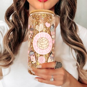 May include: A gold glitter tumbler with a pink and white design featuring the text "Cafecito Y Chismes". The tumbler has a straw and is decorated with images of coffee, pastries, and hearts.