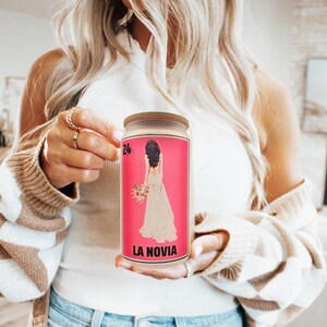 May include: A glass jar with a wooden lid. The pink label features a bride illustration and the text "LA NOVIA". The jar is held by a person wearing a white top and a beige and white striped cardigan. The number "24" is in the upper left corner.