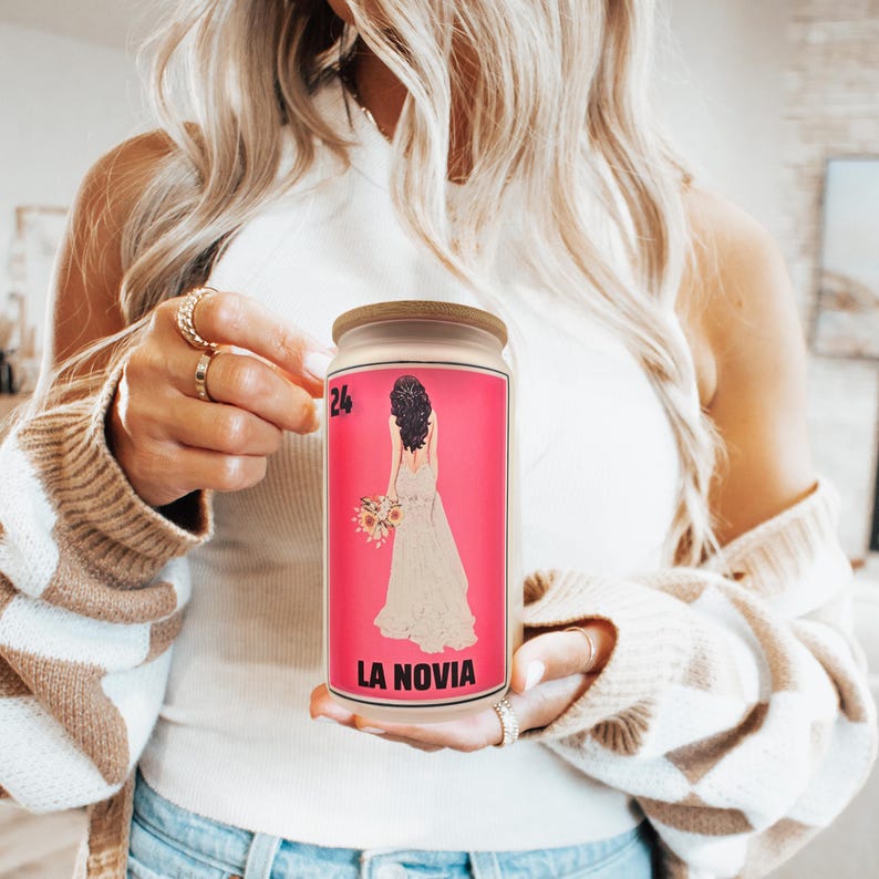 May include: A glass jar with a wooden lid. The pink label features a bride illustration and the text "LA NOVIA". The jar is held by a person wearing a white top and a beige and white striped cardigan. The number "24" is in the upper left corner.