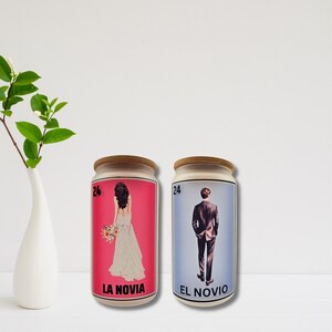 May include: Two decorative jars with wooden lids. One jar features a bride in a white wedding dress on a pink background, labelled "LA NOVIA." The other jar shows a groom in a suit on a blue background, labelled "EL NOVIO."