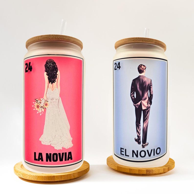 May include: Two glass can-shaped tumblers with bamboo lids and straws. One features a bride illustration on a pink background with "LA NOVIA" text. The other has a groom illustration on a blue background with "EL NOVIO" text. Both have the number 24.