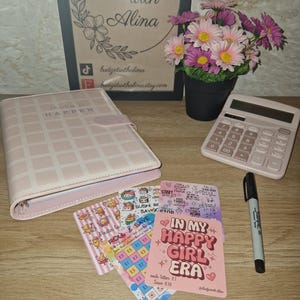May include: A selection of budgeting and planning tools. Features a pink planner with "Make it Happen" text, savings cards, a calculator, a black marker, and a framed sign that says "Budget with Alina". A small pot of pink and purple flowers is also present.