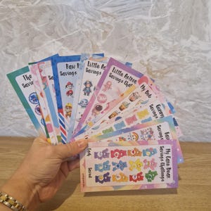 May include: A selection of colourful savings challenge cards are fanned out in a hand. The cards feature themes such as Paw Patrol, Little Astronauts, and Care Bears. Text on the cards includes "The Savir", "My Bob Savings", and "My Care Bear Savings Challenge".