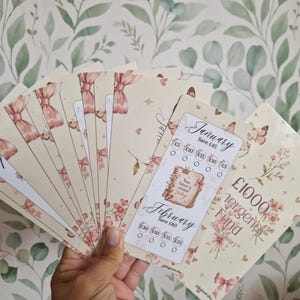 May include: A fan of rectangular paper savings trackers featuring a cream background with pink bows, flowers, and butterflies. The trackers include text for January and February, savings goals, and a section labelled £1000 Emergency Fund.