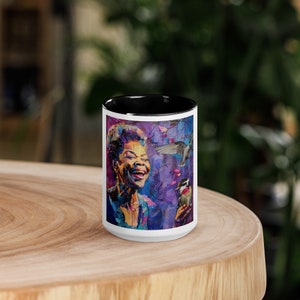 May include: A white ceramic mug with a black interior and a colorful portrait of a woman with birds and a cityscape. The artwork features vibrant blues, purples, and greens. The mug is sitting on a wooden surface.