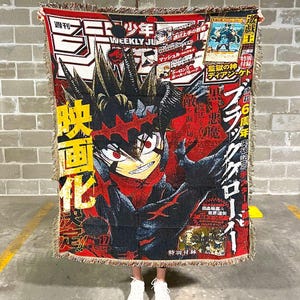 Könnte beinhalten: Eine rechteckige Decke mit einer Manga-Illustration in Rot, Schwarz und Gelb. Die Decke zeigt eine zentrale Figur und japanischen Text, darunter "WEEKLY JUMP". Sie hat Fransen an den Rändern.