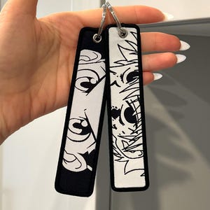 May include: Two rectangular fabric keychains featuring black and white embroidered anime-style eye designs. One keychain has a black background with white eyes, while the other has a white background with black eyes. Both include silver metal rings.