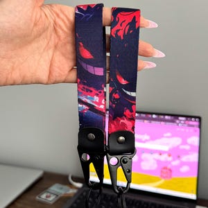 May include: Two dark blue lanyards with a colorful graphic design featuring a purple and red character. The lanyards have black metal clasps and are held up against a blurred background.
