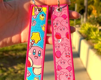 Pode incluir: Dois chaveiros retangulares com designs rosa e azul. Um apresenta um personagem de desenho animado com uma estrela e um bastão de doces em um fundo azul. O outro tem um fundo rosa com vários rostos de personagens de desenho animado. Ambos têm bordas rosa e argolas prateadas.