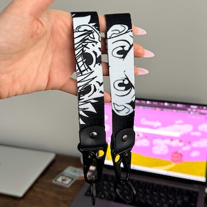 Puede incluir: Dos llaveros en blanco y negro con diseños de ojos de dibujos animados. Los llaveros tienen cierres de metal negro. Un llavero presenta un diseño abstracto en blanco y negro, el otro tiene un fondo blanco con ojos de dibujos animados.