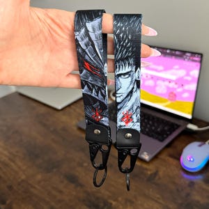 May include: Two black and white lanyards featuring detailed illustrations of a character with red accents. Each lanyard has a black clasp and a key ring. The lanyards are held up in front of a laptop.