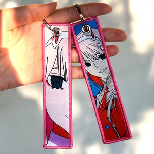 May include: Two pink and white anime character keychains with pink stitching. The keychain on the left shows a close-up of the character's eye. The keychain on the right shows the character's full face and body.