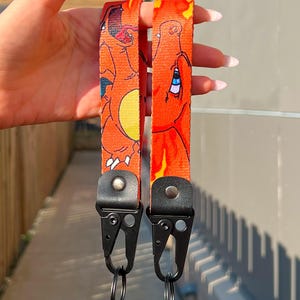 Puede incluir: Dos cordones naranjas con un diseño de personaje de dibujos animados. Los cordones tienen cierres y llaveros de metal negro. El personaje es naranja con detalles amarillos y un ojo azul.