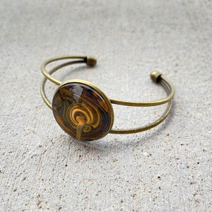 May include: A gold-toned metal bangle bracelet with a round, brown and gold resin cabochon. The cabochon has a swirling pattern.