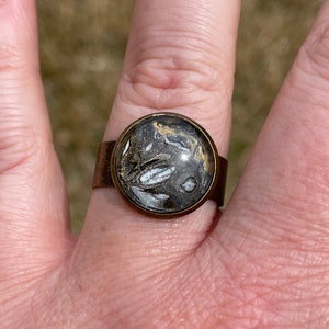 May include: A brass ring with a round, black, white, and gold resin inlay. The resin has a swirling, abstract pattern.