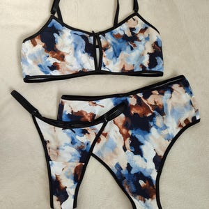 Op de afbeelding: Een tweedelige lingerieset met een aquarelpatroon in blauw, bruin en wit. De set bevat een bralette met verstelbare bandjes en een sleutelgatdetail, en twee bijpassende slips met zwarte afwerking.