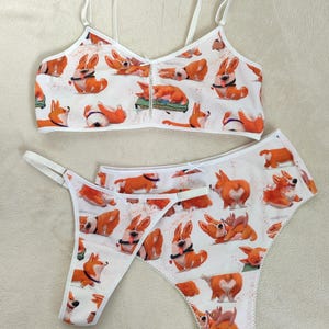 Op de afbeelding: Witte lingerie set met een patroon van oranje en witte corgi honden. De set bestaat uit een bralette met dunne bandjes en een string. De randen zijn afgewerkt met witte kant.