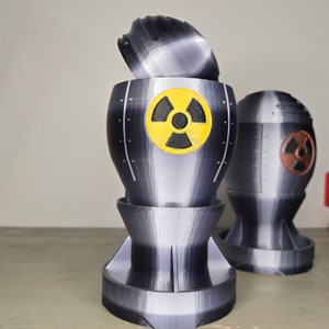 May include: A gray and black 3D-printed bomb-shaped container with a yellow radiation symbol. The container has a removable top and is cylindrical with a flared base. Another similar container is visible in the background.