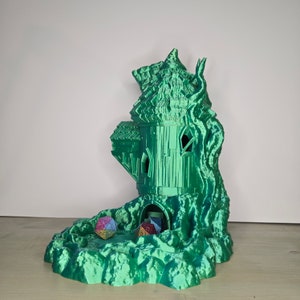 May include: A green 3D printed dice tower shaped like a treehouse with a small opening at the bottom. Two multi-colored dice are visible inside the tower.