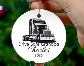 May include: A white ceramic ornament with a black and silver semi-truck illustration. The text "Drive Safe Grandpa Charles 2025" is printed below the truck. A gold string is attached for hanging.