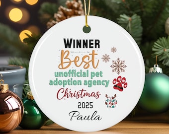 May include: A white ceramic Christmas ornament with gold string. The ornament features the words "WINNER Best unofficial pet adoption agency Christmas 2025 Paula" in various colors and fonts, along with paw prints and snowflakes.