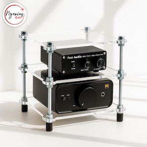 May include: A clear acrylic audio equipment stand with two black audio components. The stand is held together with silver bolts and rods. The top component has the text "Fost Audio" and the bottom component has a large knob.