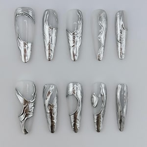 May include: Set of ten long, white, oval-shaped fake nails with silver foil accents. The foil has a textured, marbled look.