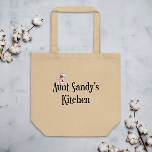 Può includere: Una borsa tote beige con una grafica nera di un pollo chef di cartoni animati e il testo "Aunt Sandy's Kitchen".