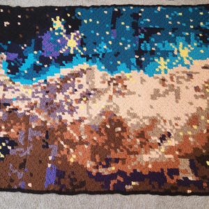 May include: A handmade, pixelated blanket with a celestial design. The blanket features a dark blue and black background with patches of brown, tan, blue, and yellow, creating a starry night effect. The blanket is rectangular.