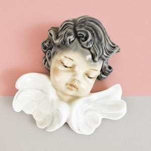 Può includere: Un angelo in ceramica, decorazione murale, con gli occhi chiusi e un'espressione serena. L'angelo ha i capelli grigi e le ali bianche. Lo sfondo è diviso in sezioni rosa e grigie.