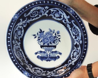 May include: A large, round, blue and white decorative plate. The plate features a floral design in the center and an ornate border. The plate is held by two hands, showcasing the intricate details and the size of the plate.