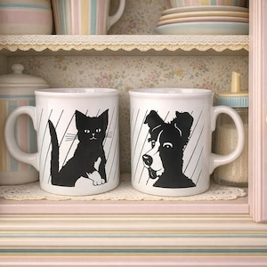 May include: Two white ceramic mugs with black cat and dog silhouettes. The mugs are on a shelf inside a pastel-colored cabinet. Other mugs and dishes are visible on the shelves above.