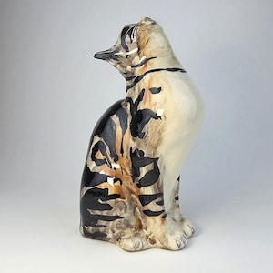 Könnte beinhalten: Eine Keramik-Katzenfigur in sitzender Pose, mit cremefarbenem Körper und schwarz-braunen tigerähnlichen Streifen. Die Katze hat einen langen, schlanken Hals und blickt nach oben. Die Figur steht vor einem weißen Hintergrund.