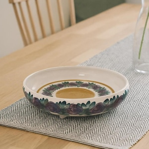 May include: A white ceramic serving bowl with a floral design in purple, green, and gold. The bowl sits on a gray and white herringbone patterned placemat on a wooden table. A wooden chair is in the background.