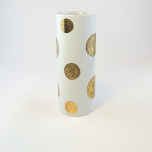 Pode incluir: Um vaso cilíndrico branco adornado com desenhos de moedas douradas. O vaso tem uma superfície lisa e um design simples e elegante. As moedas de ouro são de vários tamanhos e estão uniformemente espaçadas.