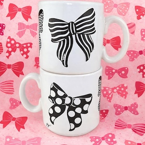 May include: Two white ceramic mugs stacked on top of each other, each with a black bow design. One bow is striped, the other is polka-dotted. The background is pink with various bow patterns in shades of pink and red.