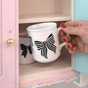 May include: White ceramic mug featuring a black bow design. The mug is held in a pink cabinet with another mug on the shelf. The cabinet has a gold knob and a scalloped trim. The background is light blue and pink.