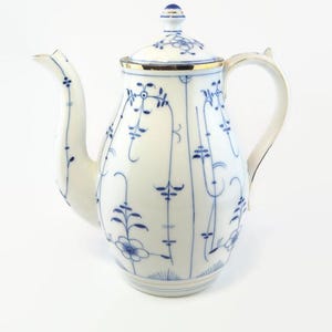 Puede incluir: Una cafetera de porcelana blanca con un patrón floral azul y blanco y ribete dorado. La cafetera tiene un cuerpo redondo y un pico en el lateral. La tapa tiene un pomo en la parte superior.