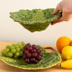 Könnte beinhalten: Eine grüne Keramik-Servierschüssel und -platte, beide blattförmig, mit Trauben und anderen Früchten. Die Schüssel wird von einer Hand gehalten. Weitere Früchte sind eine Orange, ein Apfel, eine Zitrone und eine Banane.