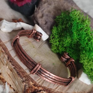 Puede incluir: Una pulsera de puño de color cobre con un diseño en capas. La pulsera está hecha de múltiples alambres paralelos, sujetos por bandas más gruesas. Los extremos de la pulsera tienen una apariencia tejida. La pulsera se exhibe sobre un trozo de madera.