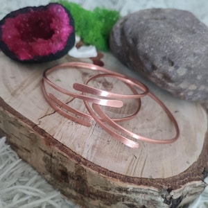 Puede incluir: Brazaletes de metal color cobre con un diseño abierto se exhiben sobre una rodaja de madera. Los brazaletes tienen un estilo minimalista. Una geoda con interior rosa, musgo verde y una piedra gris también están en la imagen.